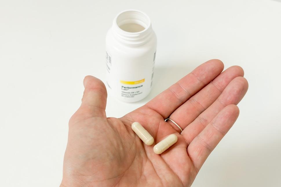 Closeup of an open palm holding two beige capsules of Performance Lab NutriGenesis D3 + K2. You can see the open pill bottle on the white countertop below.