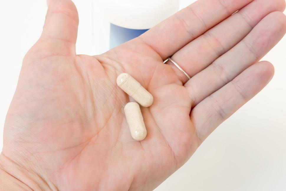 An open palm with two off-white capsules of Performance Lab NutriGenesis Multi for Men. You can see the pill bottle below on the white countertop.