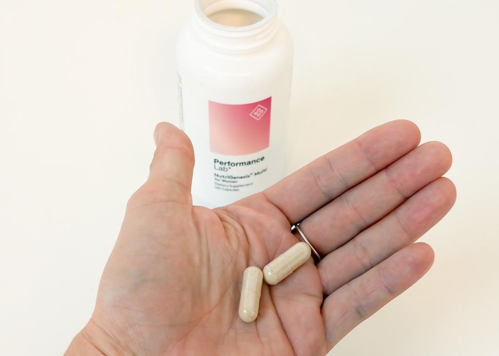 An open palm holding two beige-colored Performance Lab NutriGenesis Multi for Women capsules. You can see the open pill bottle on a white countertop below.