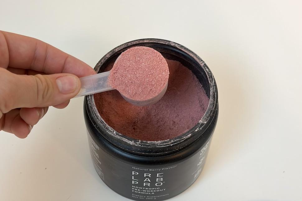 An open canister of Performance Lab Pre Lab Pro. The canister is black and the powder inside is a light purple. Someone is scooping out the powder with a plastic scooper.