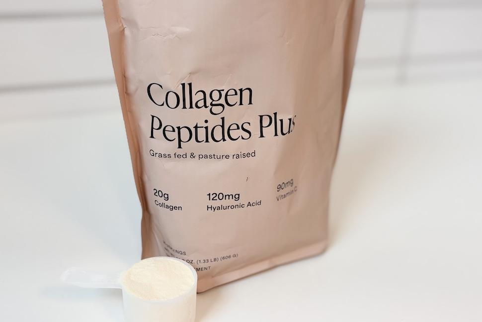 A brown bag of Collagen Peptides Plus sitting on a white countertop. Next to it is a plastic scooper packed to the brim with the off-white powder.