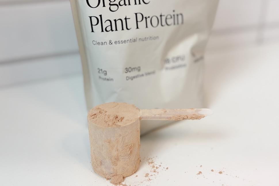 A bag of Quince Organic Plant Protein sitting on a white countertop. A scooper packed to the brim with the light-brown powder sits next to it.