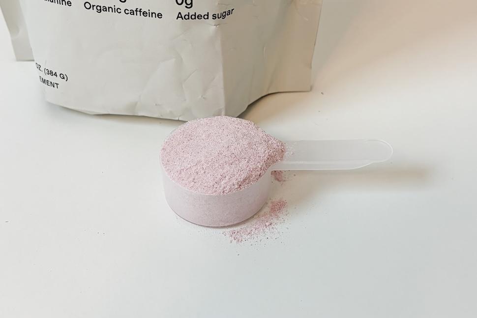 Closeup shot of a big of Quince Performance Pre Workout sitting on a white countertop. A plastic scooper packed with the faint purple powder sits next to it.