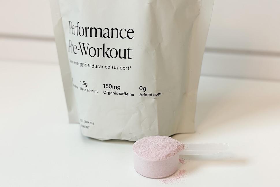 A big of Quince Performance Pre Workout sitting on a white countertop. A plastic scooper packed with the faint purple powder sits next to it.