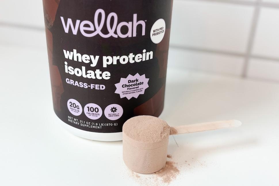 Wellah Grass-Fed Whey Protein + Pre and Probiotics sitting on a white countertop. A scoop packed with light-brown powder sits next to it.