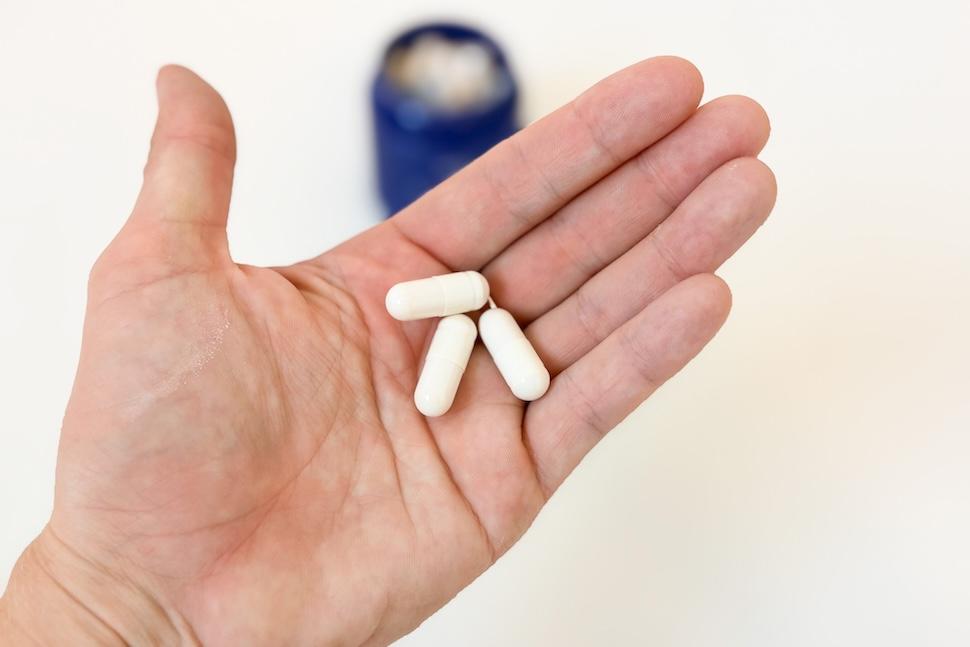 Closeup of a hand holding three off-white Welzo Akkermansia capsules. You can see an open bottle of the bills on the white countertop below.