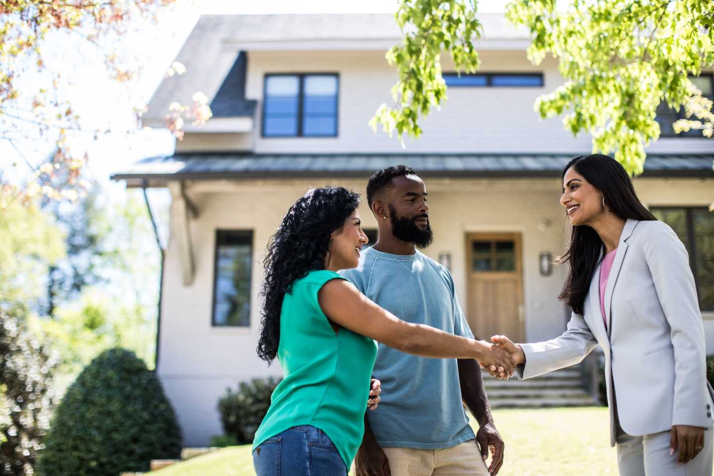 Two women, with a man standing next to them, shake hand in front of a house.