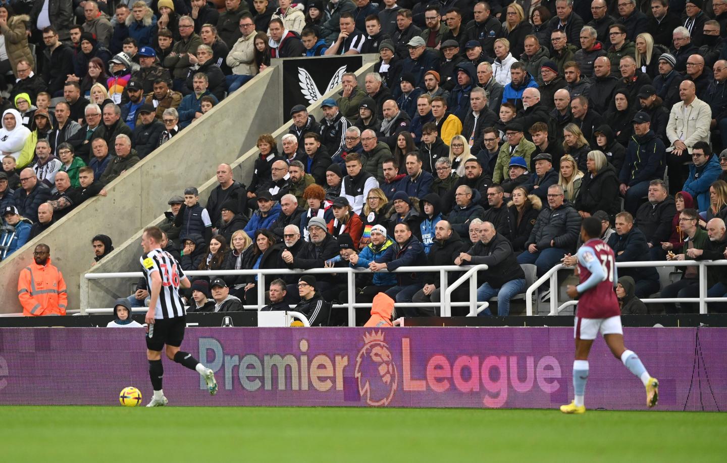 Two soccer players run across the field in front of bleachers filled with fans and a Premier League advertisement.