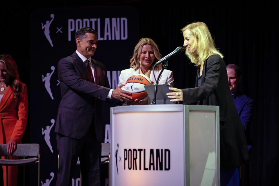 Alex Bhathal and Lisa Bhathal Merage stand next to a podium and accept a basketball from Cathy Engelbert.