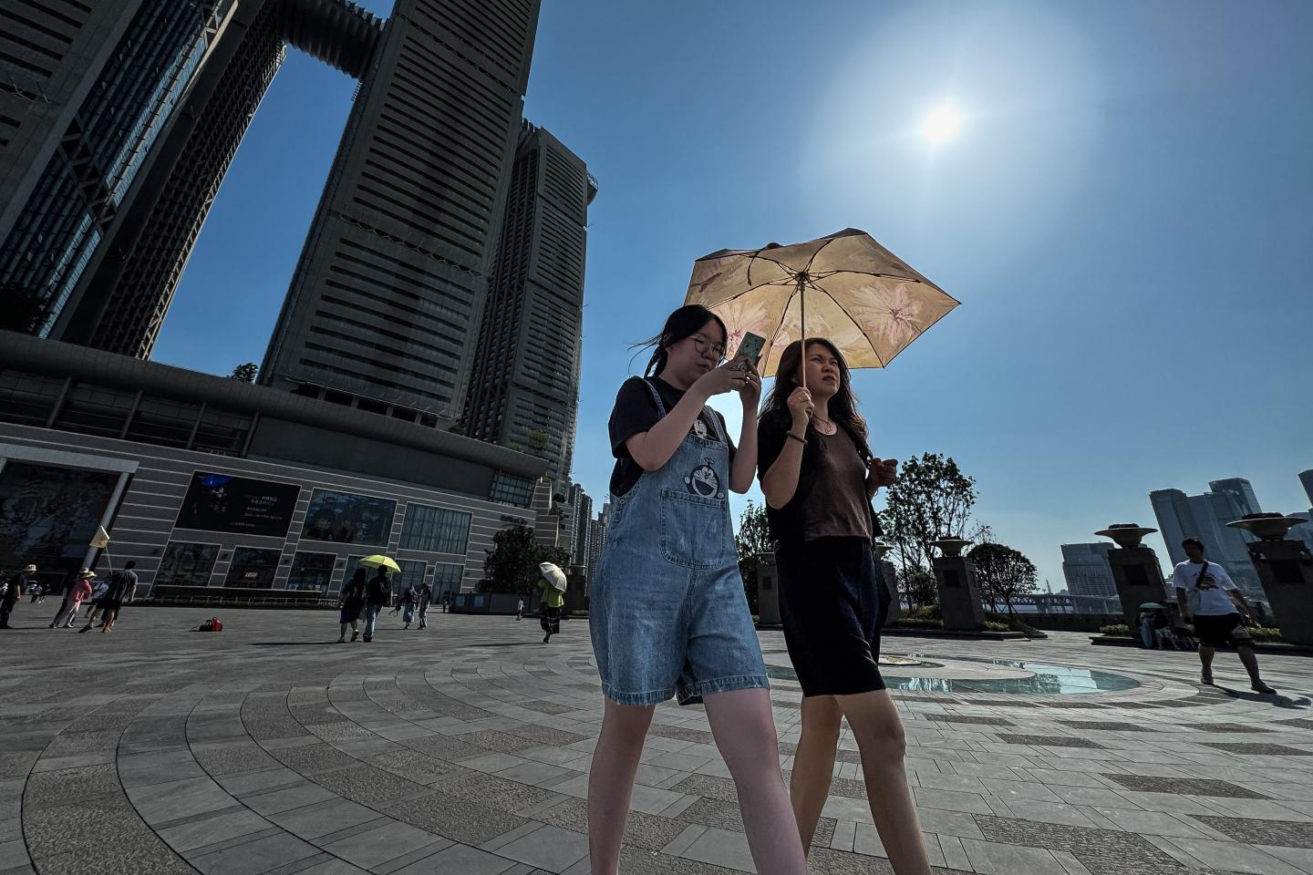 Two women share a floral-patterned sun umbrella while walking through Chaotianmen Square under the blazing sun, with the Raffles City Chongqing towers visible in the background during an extremely hot summer day, on June 16, 2025 in Chongqing, China. 