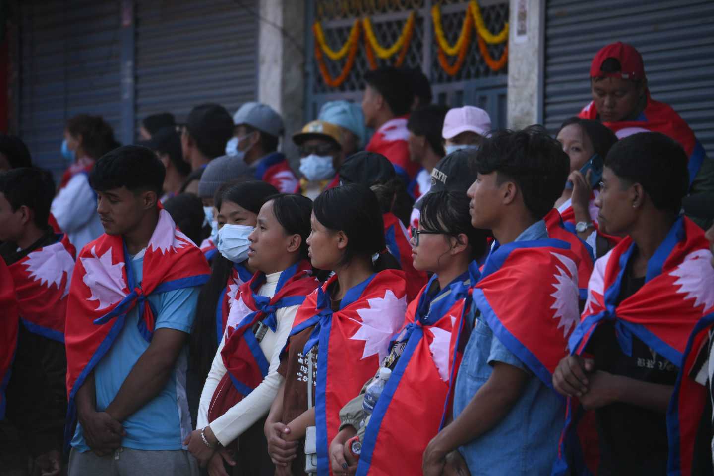 A group of young people stand together, each with the flag of Nepal draped around their shoulders.
