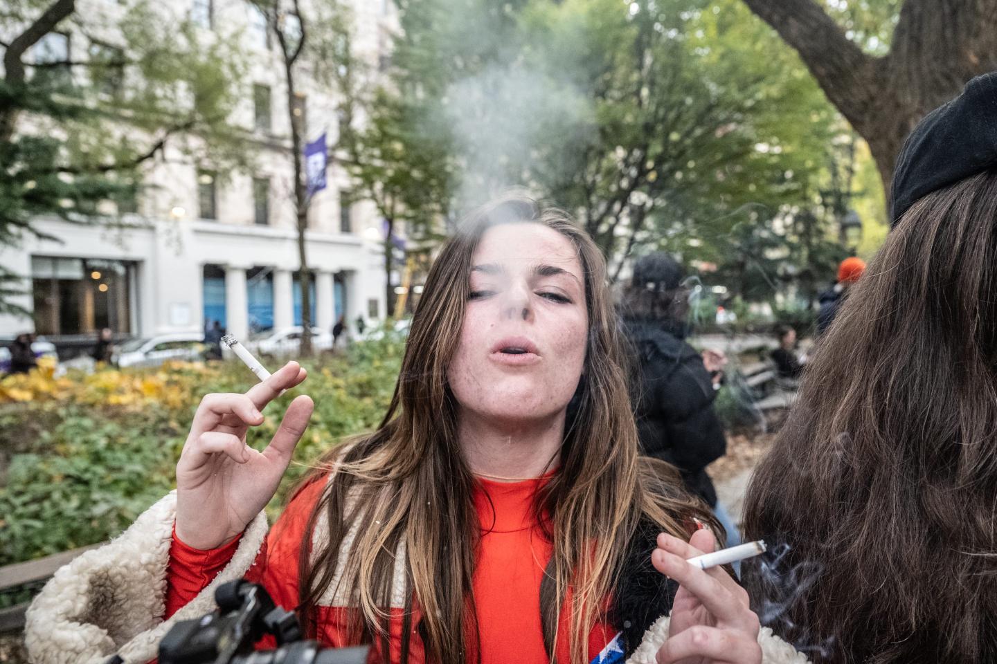 A woman in the park holds a cigarette in each hand and exhales a puff of smoke.
