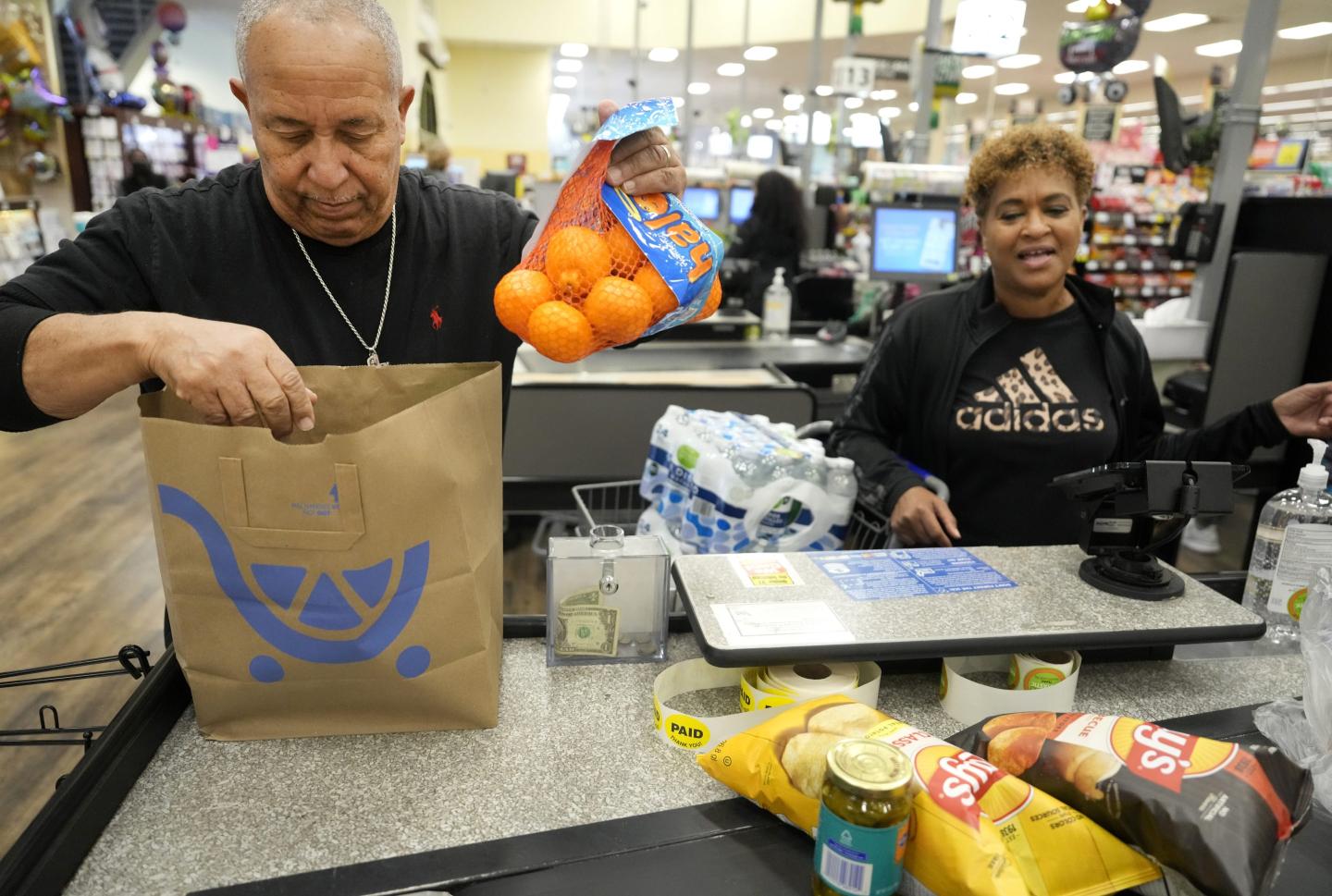 A man bags groceries in the check-out line, and a woman stands next to him.