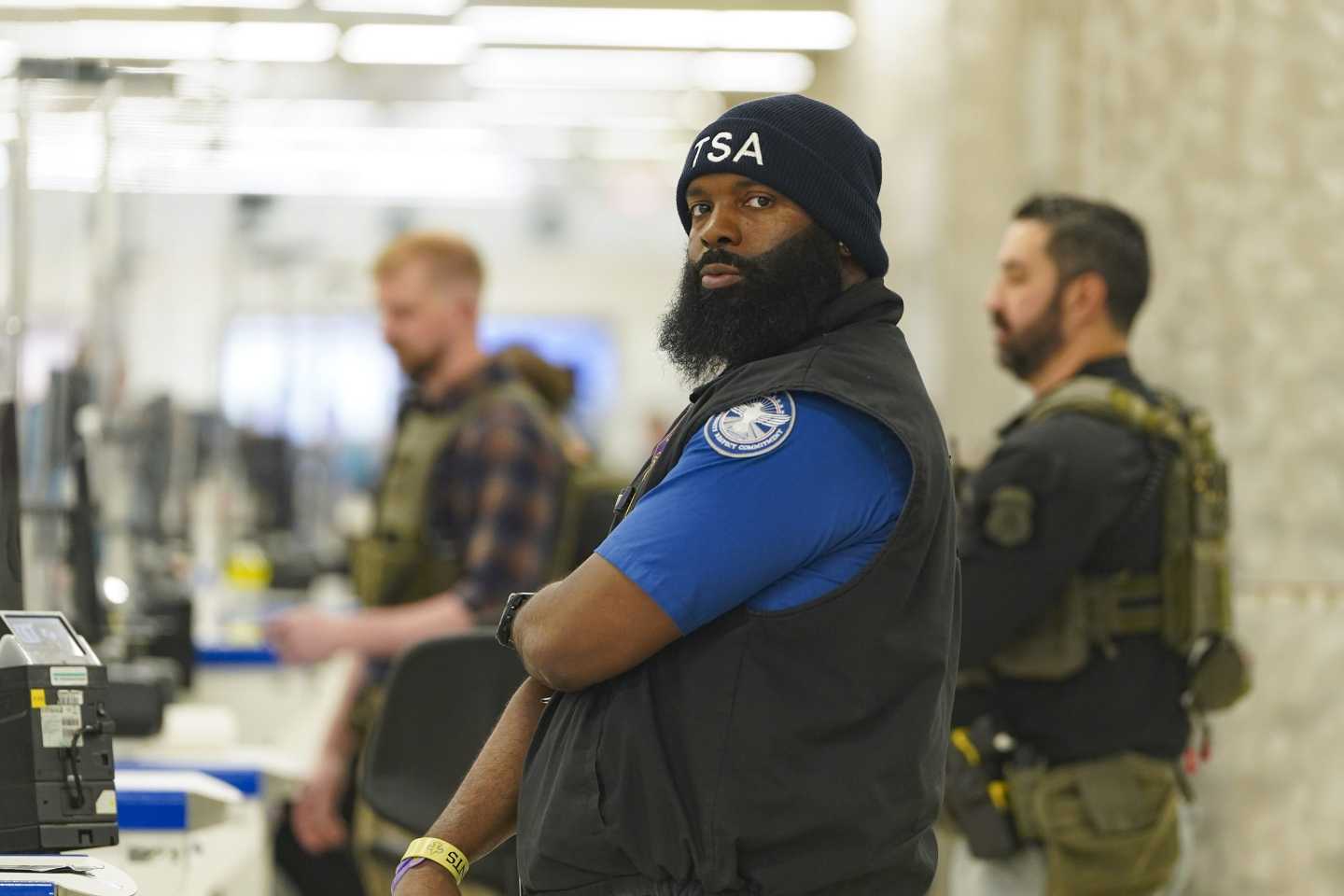 A TSA officer, with one arm crossed over his body, looks into the camera.