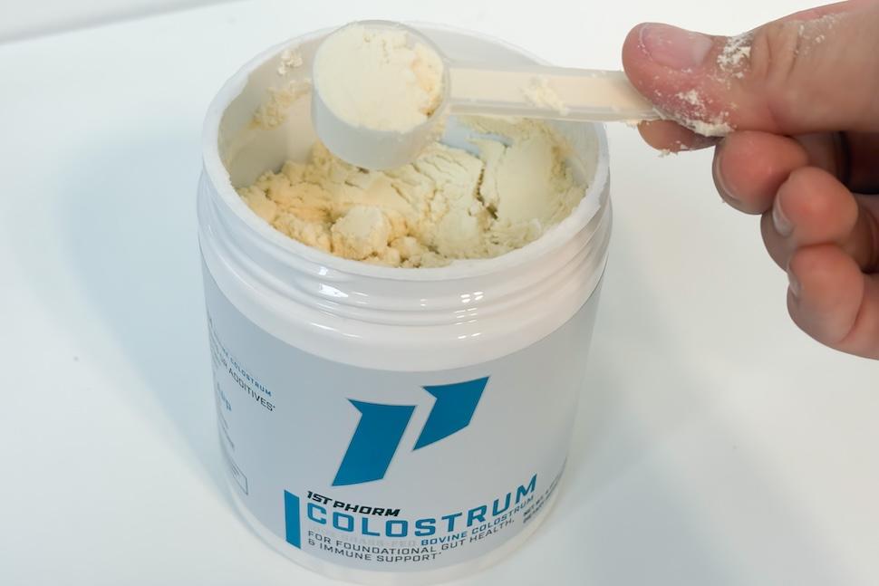 Closeup of a hand using a plastic scoop to scoop out 1st Phorm Colostrum from its canister. The powder is off-white in color.