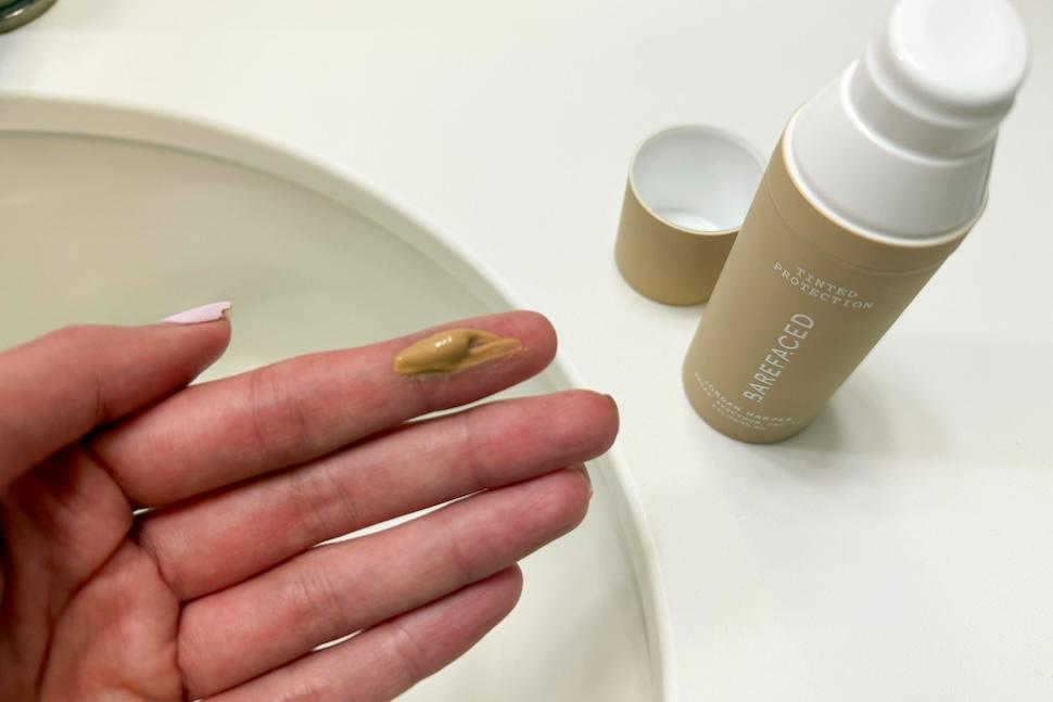 Closeup of a hand with one dab of Barefaced Tinted Protection SPF 50+ on the index finger. You can see the bottle of moisturizer on the white countertop below.