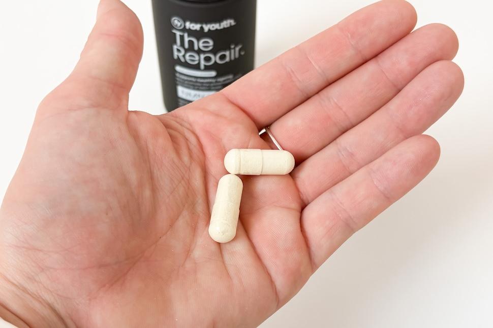 Closeup of an open palm holding two off-white capsules of For Youth The Repair NMN+. You can see the black bottle of the supplement on the white countertop below.