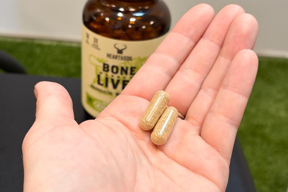 An open palm holding two capsules of Heart and Soil Bone Marrow and Liver capsules. You can see the bottle of the supplements on the countertop below.
