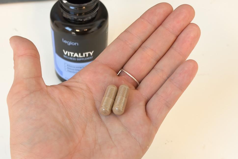 Closeup of an open palm holding two gold-ish capsules of Legion Vitality. You can see an open bottle of the capsules on the white countertop below.