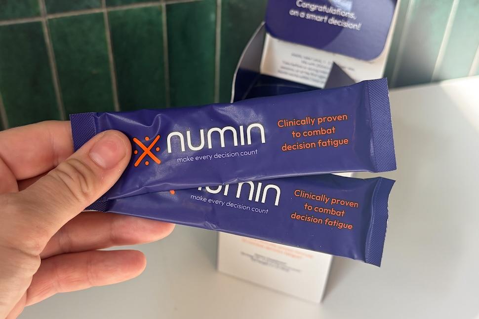 Closeup of a hand holding two individual Numin packets. You can see the box on the countertop below.