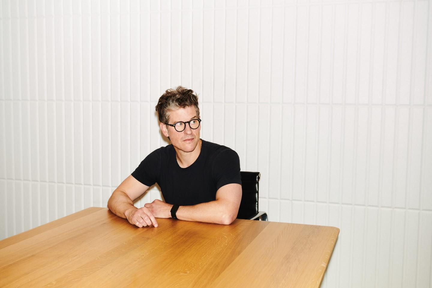 Fragment CEO sitting down with arms resting on a wooden table as he looks to his left.
