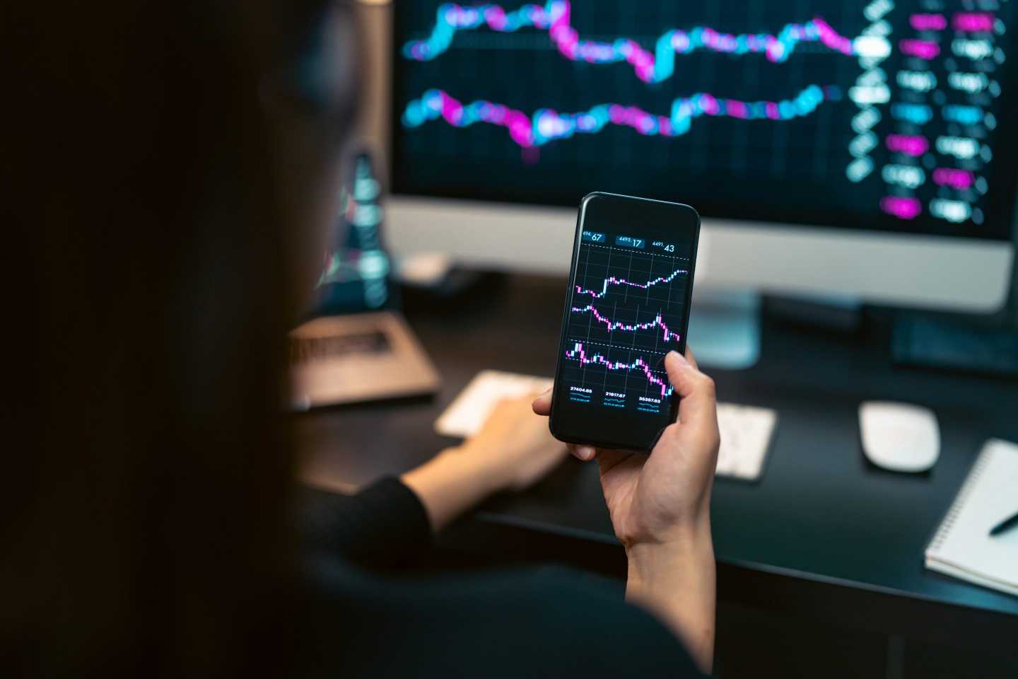 A woman trades on her phone in front of a computer with a display of crypto charts.