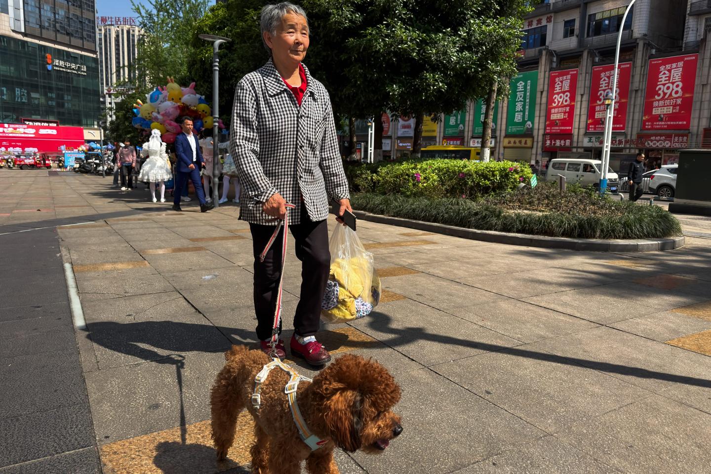 An elderly woman walks a dog while carrying shopping bags along a street on April 4, 2026, in Chongqing, China.