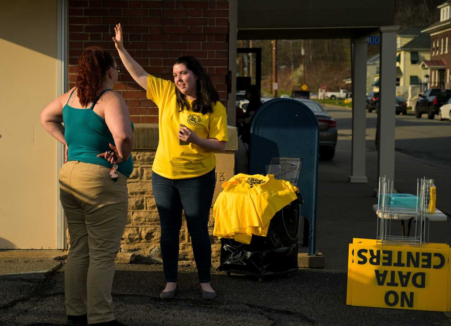 A woman in a yellow jacket in front of a "NO DATA CENTERS" sign, speaks with another woman with her hands on her hips.