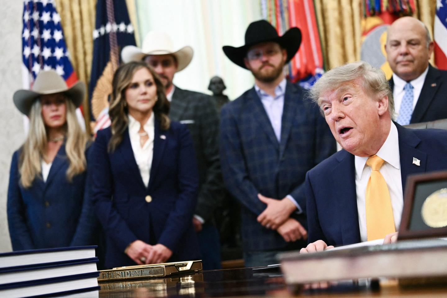 Donald Trump sits in the Oval Office with people in cowboy hats standing behind him.