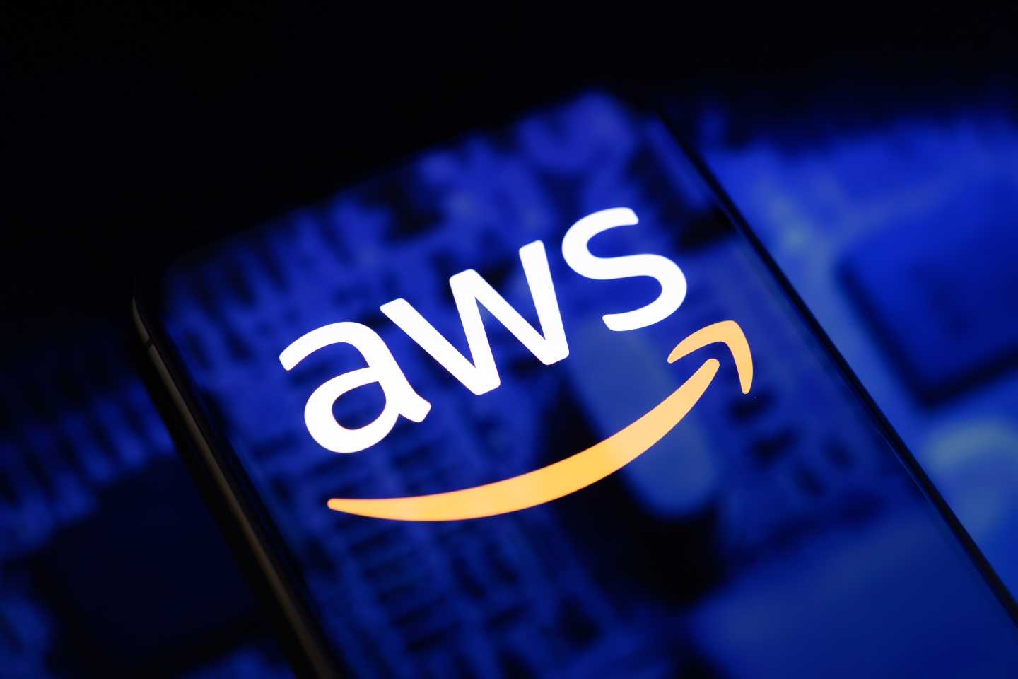 The AWS logo is displayed on a smartphone screen placed on a reflective surface onto which an illustration of blue printed circuit boards.