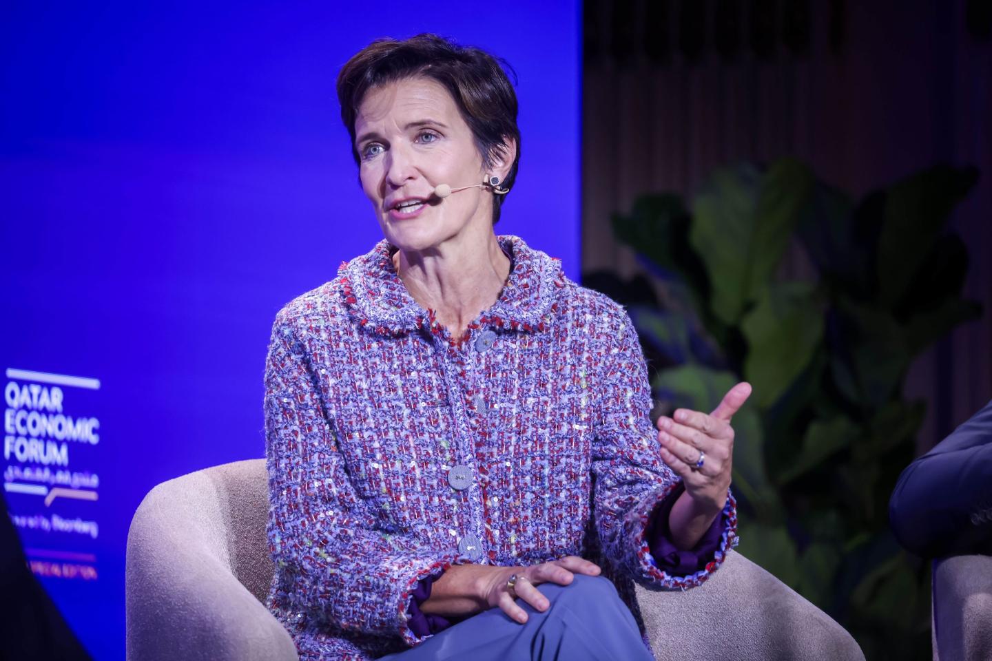 Jane Fraser, sitting on a light-colored chair onstage, speaks and gestures with her hand.