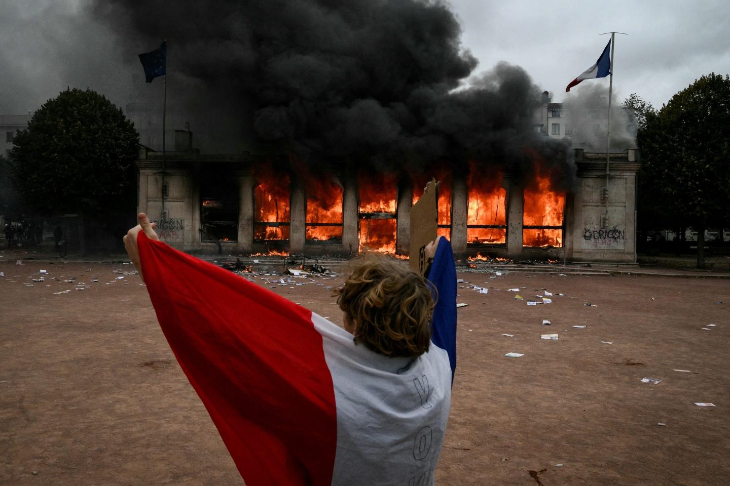 A protester wrapped in a French national flag stands in front of Lyon's tourism office set on fire during a demonstration to protest against the high cost of living on October 17, 2026.
