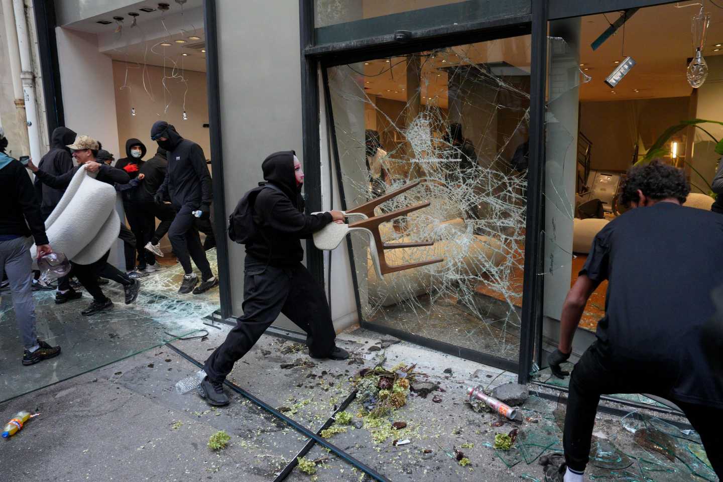 A person wearing all black throws a chair at a glass window in the midst of a riot.