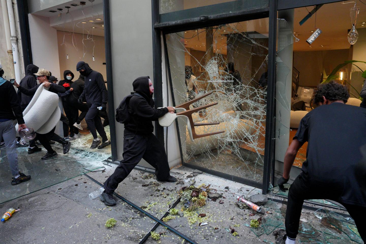 A person wearing all black throws a chair at a glass window in the midst of a riot.